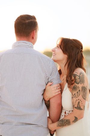Smiling tattooed woman in white dress embraces man in striped shirt outdoors at golden hour.