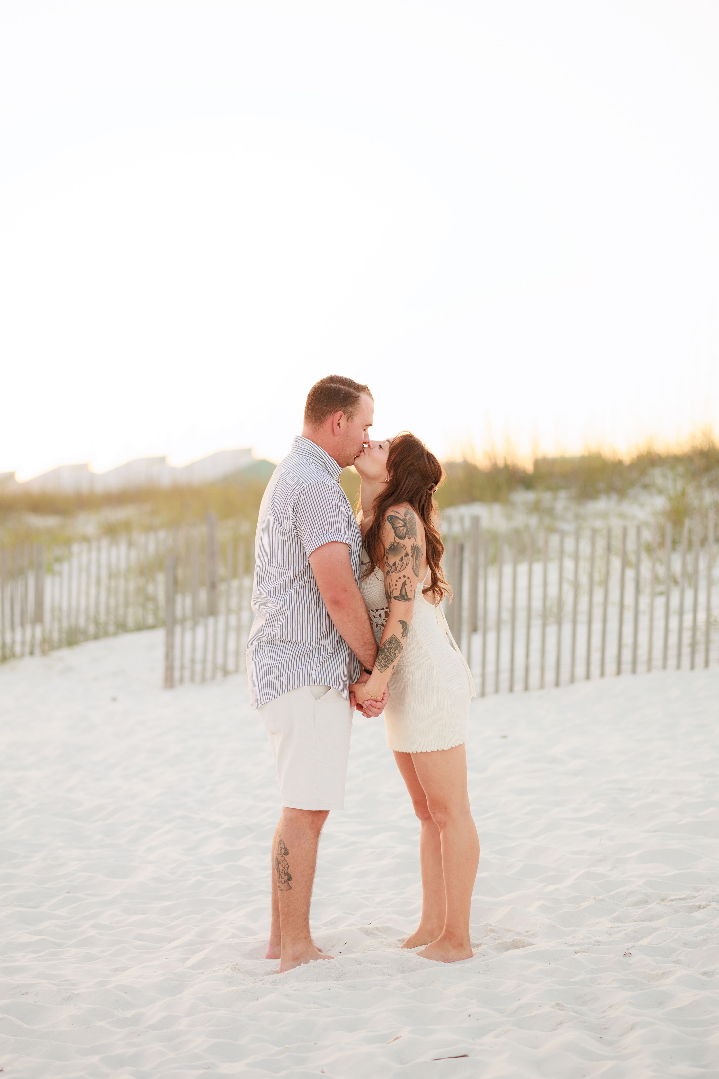 Couple kissing on white sand beach at sunset, woman with tattoos in white dress holding hands with man in blue shirt.