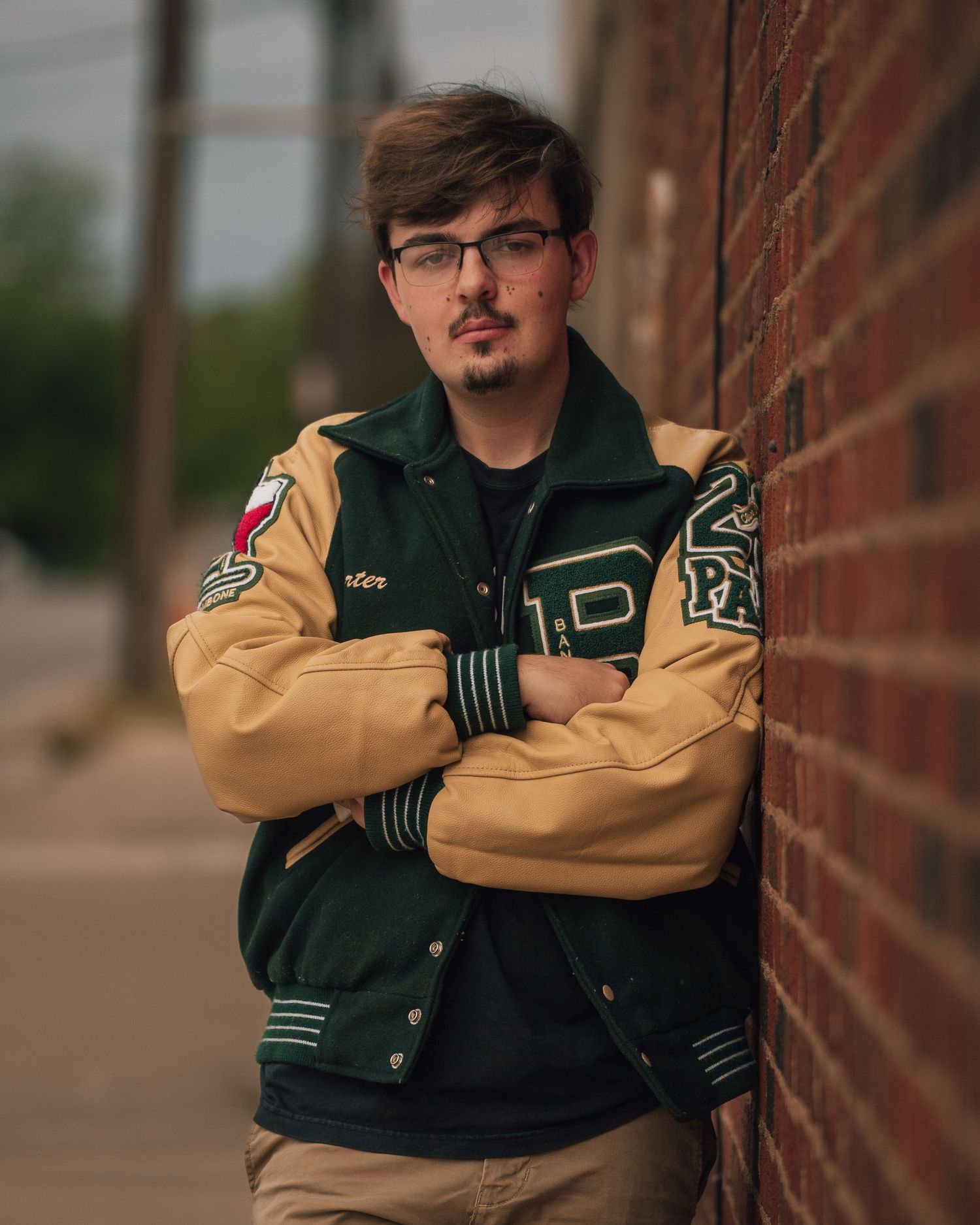 Young man in green and gold varsity jacket leaning against a brick wall with arms crossed outdoors.