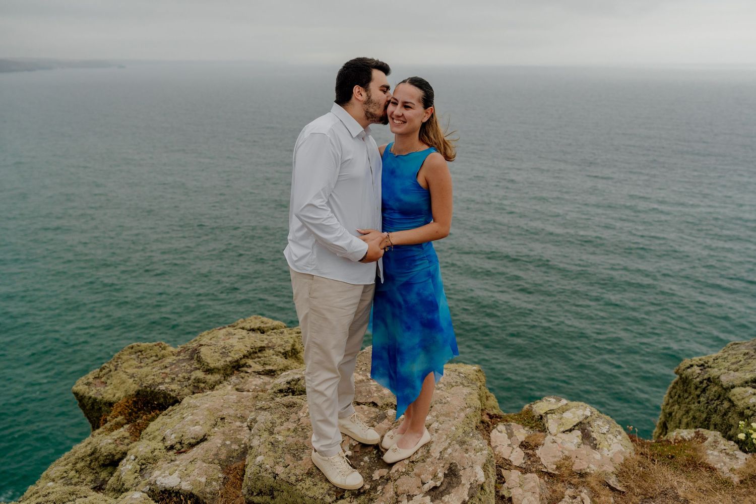 Couple embracing on st agnes head overlooking turquoise ocean, woman in blue dress, man in white outfit.