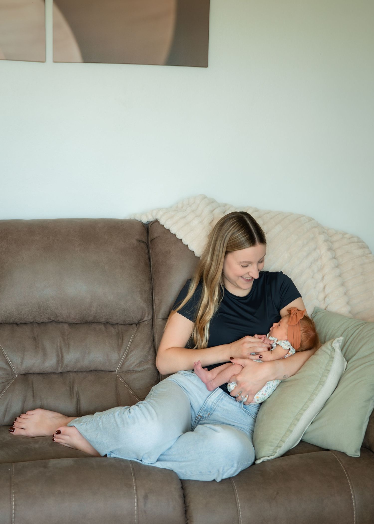 A young mother sits on a brown sofa cradling her newborn baby, smiling warmly in a cozy living room.