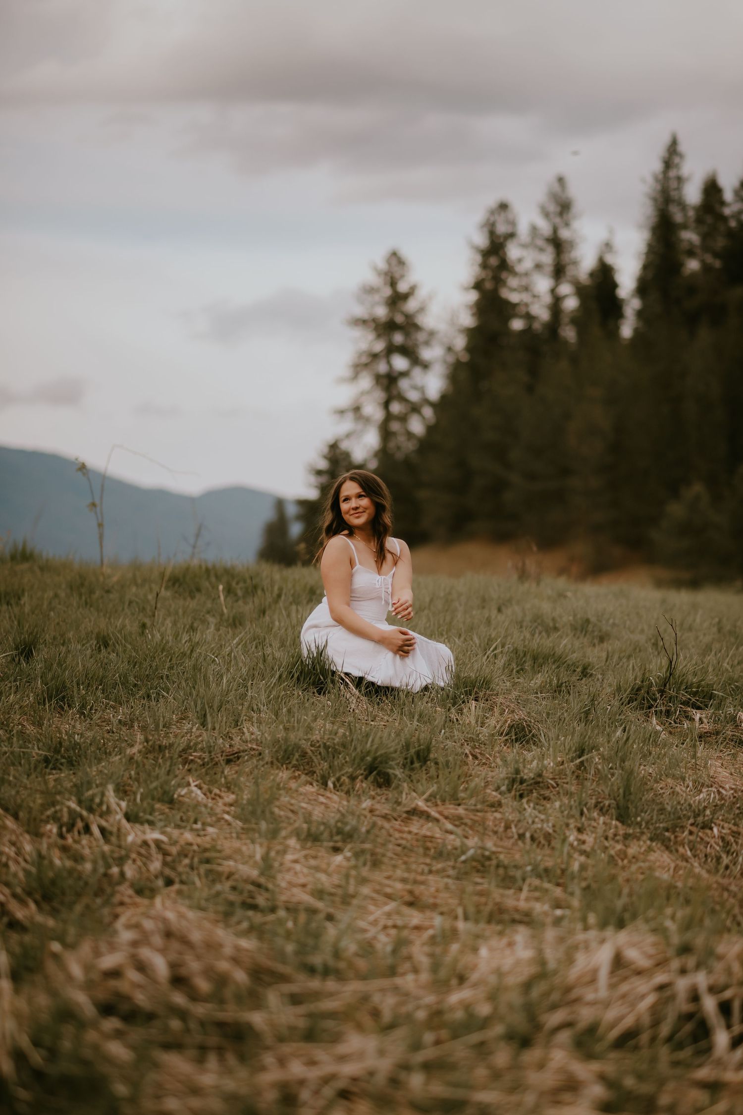 Pregnant woman in white dress sitting in a mountain meadow surrounded by evergreen trees and overcast skies.