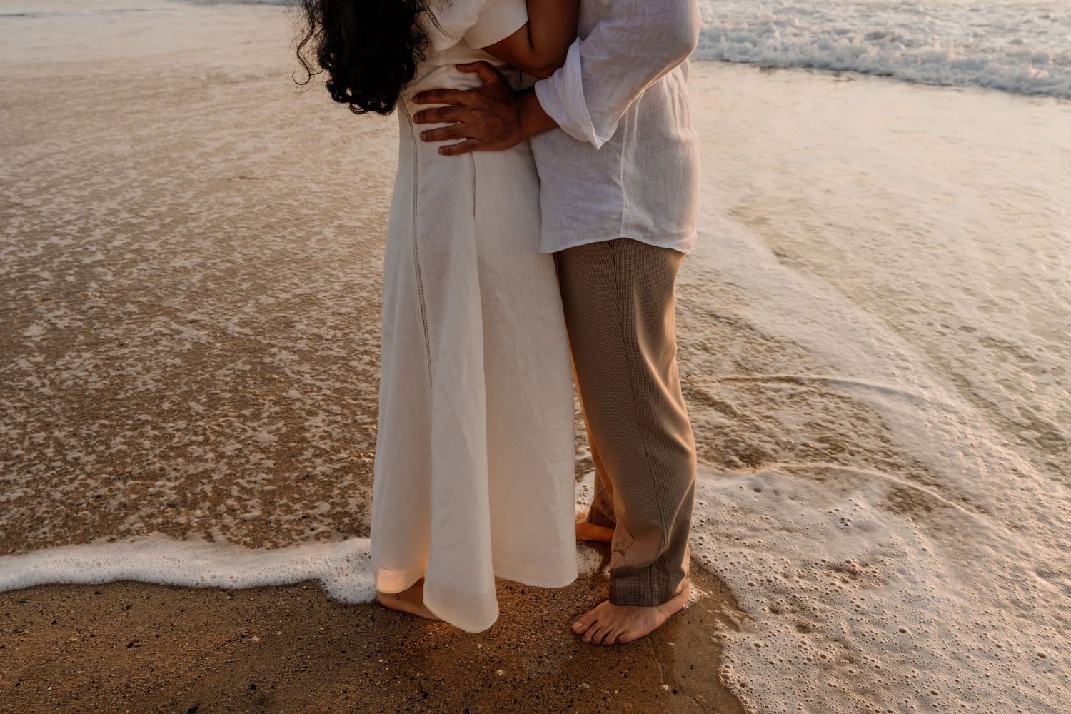 Couple embracing barefoot on sandy beach shore at sunset, woman in white dress, man in linen shirt and khaki pants.