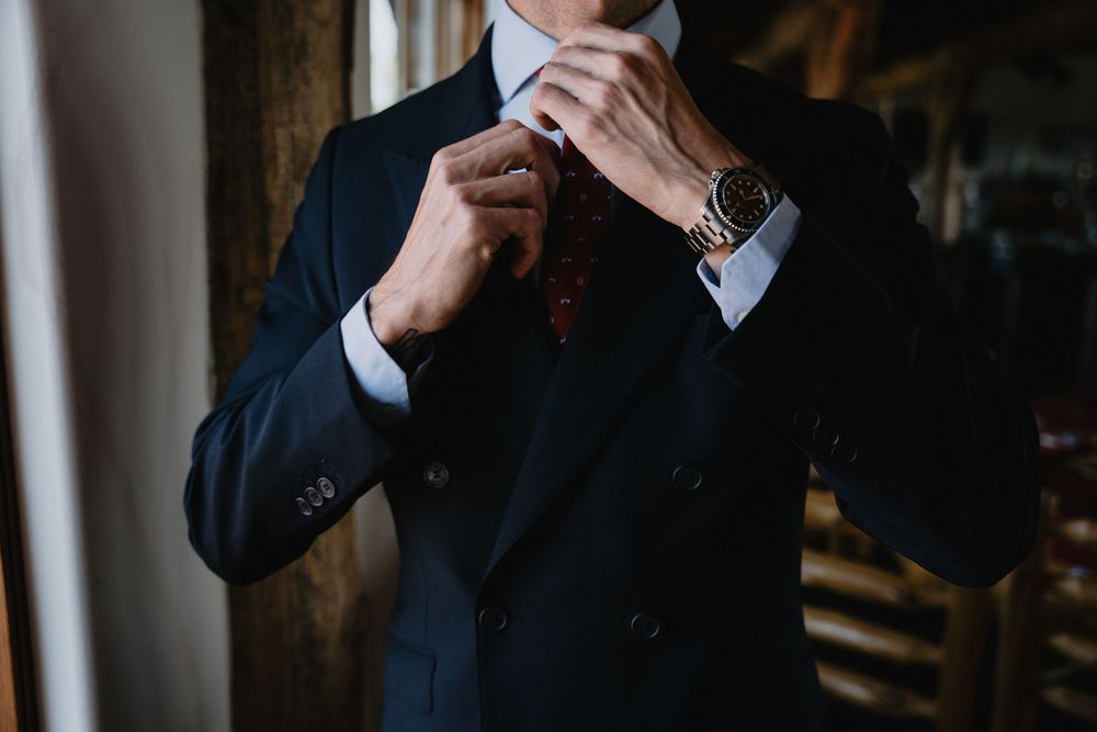 Man in navy double-breasted suit adjusting red floral tie, wearing gold watch, elegant formal wedding attire.