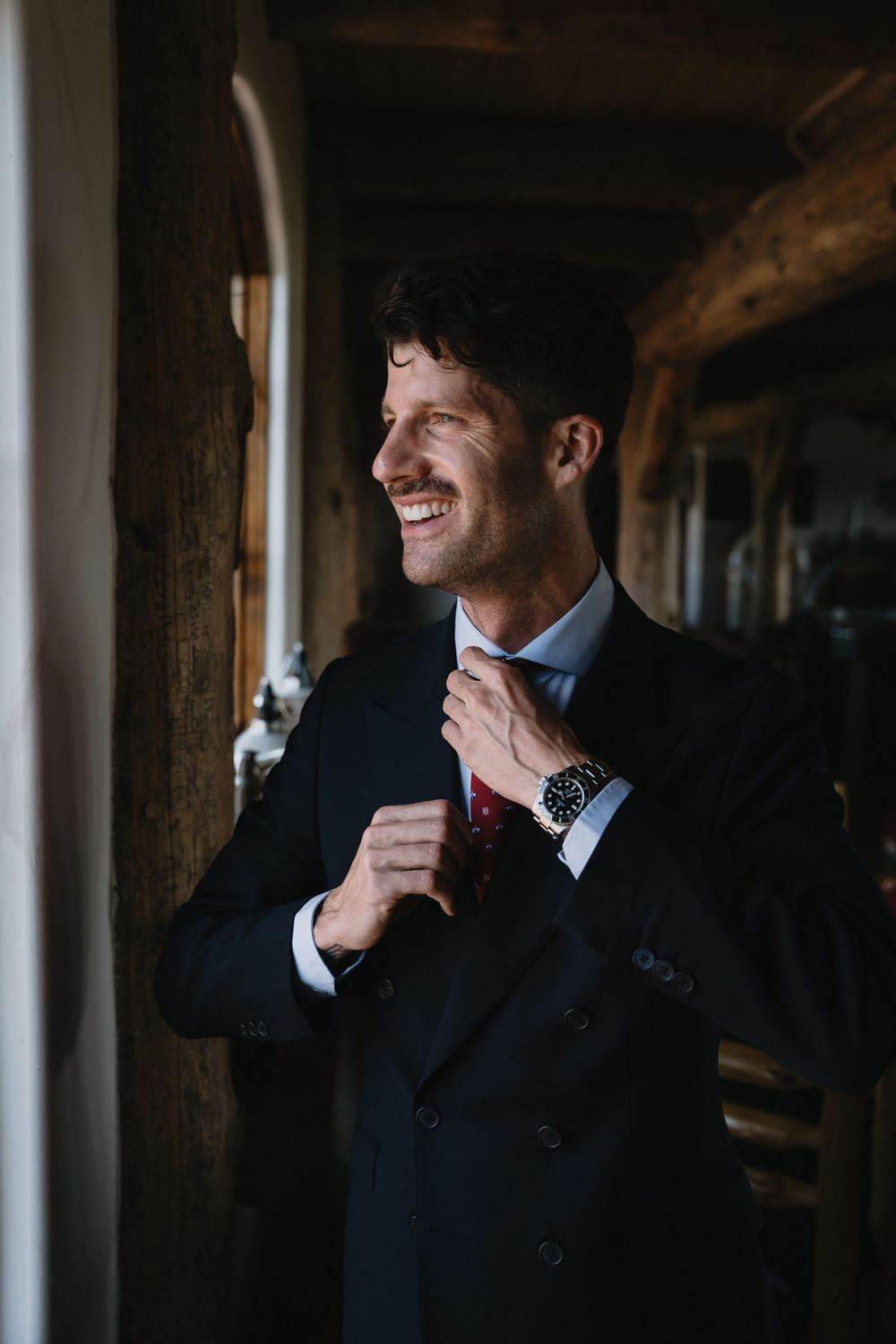 Smiling groom in dark suit adjusting red tie, wearing a watch, standing in rustic wooden doorway.
