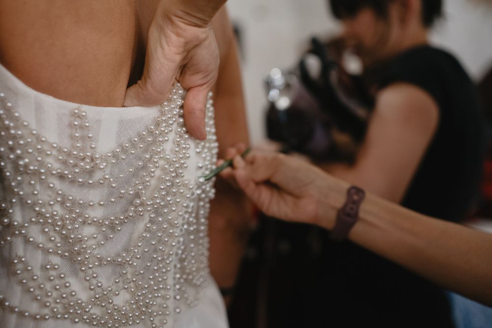A wedding dress adorned with pearl details being fastened by a helper during bridal preparations.