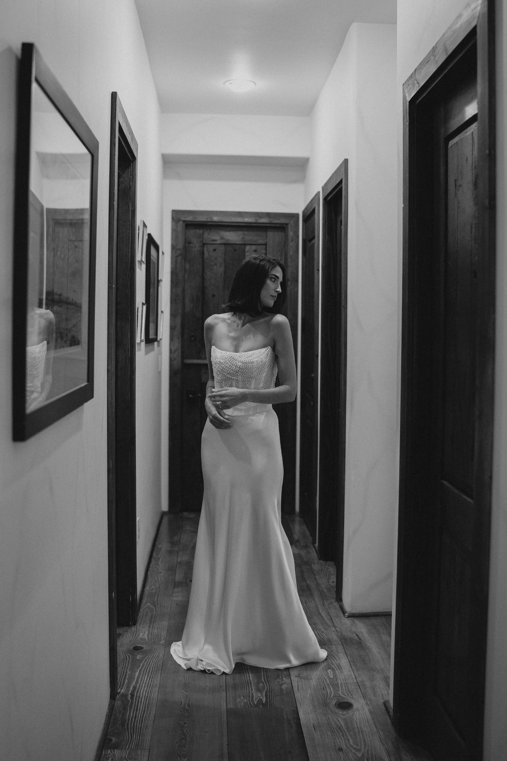 Black and white photo of a bride in a strapless wedding gown standing in a narrow hallway with wooden floors.