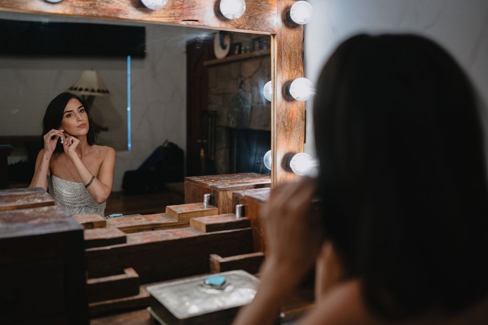 Woman adjusting earrings at a Hollywood vanity mirror with bright bulb lights in a dressing room.