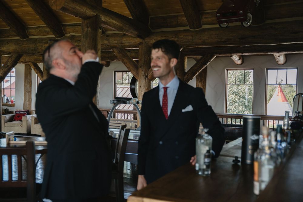 Two men in suits share a laugh at a rustic bar venue, one drinking while the other smiles confidently.