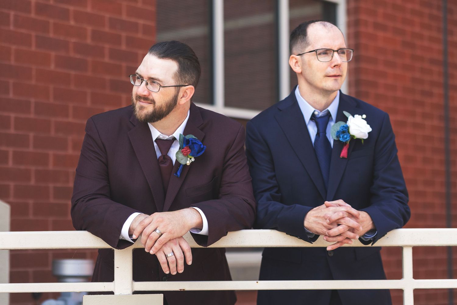Two grooms in suits with boutonnieres stand at a railing outside a brick building on their wedding day.