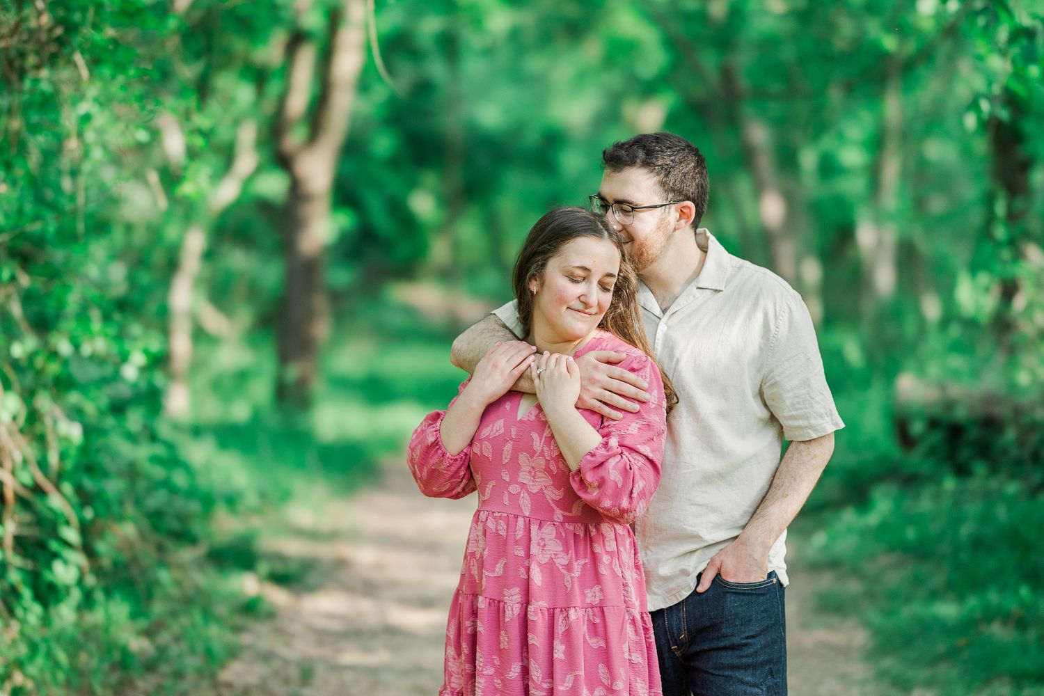 Couple embracing on a forest path, woman in pink floral dress, man in beige shirt, surrounded by lush green trees.