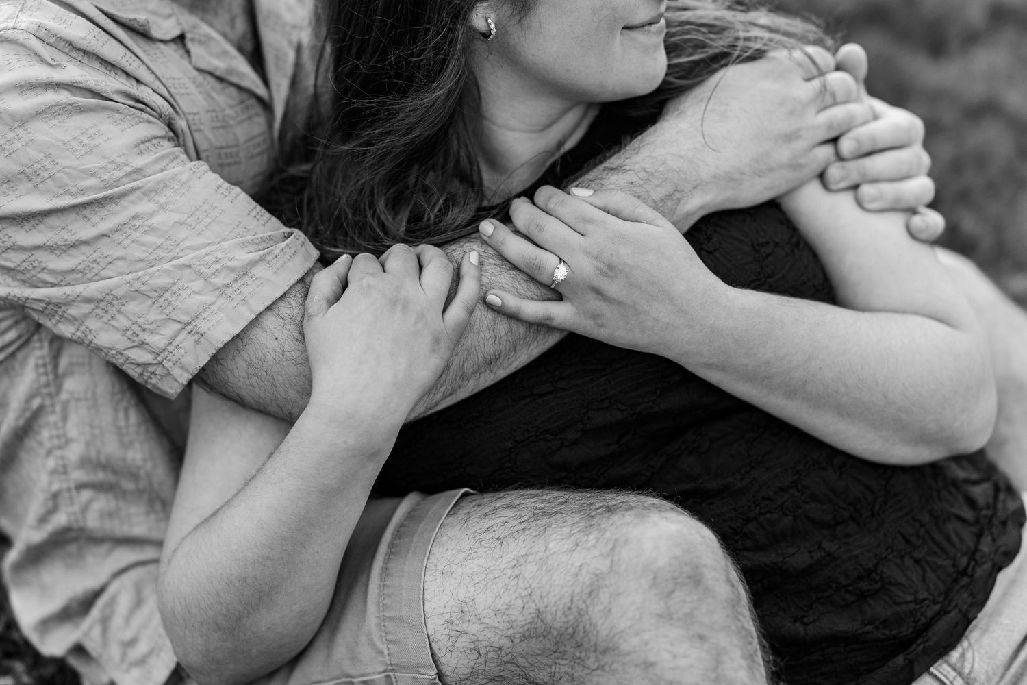 Black and white photo of a couple embracing tenderly, woman wearing engagement ring, sitting outdoors.