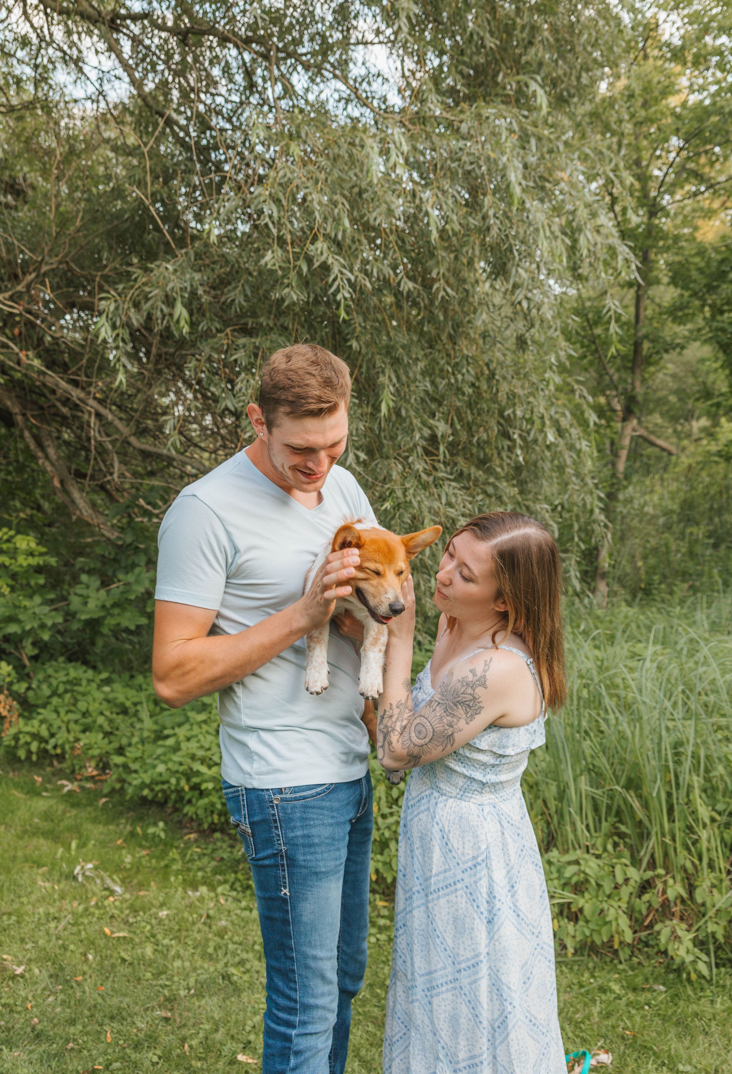 A couple smiles while holding an adorable brown and white puppy outdoors in a lush green park setting.