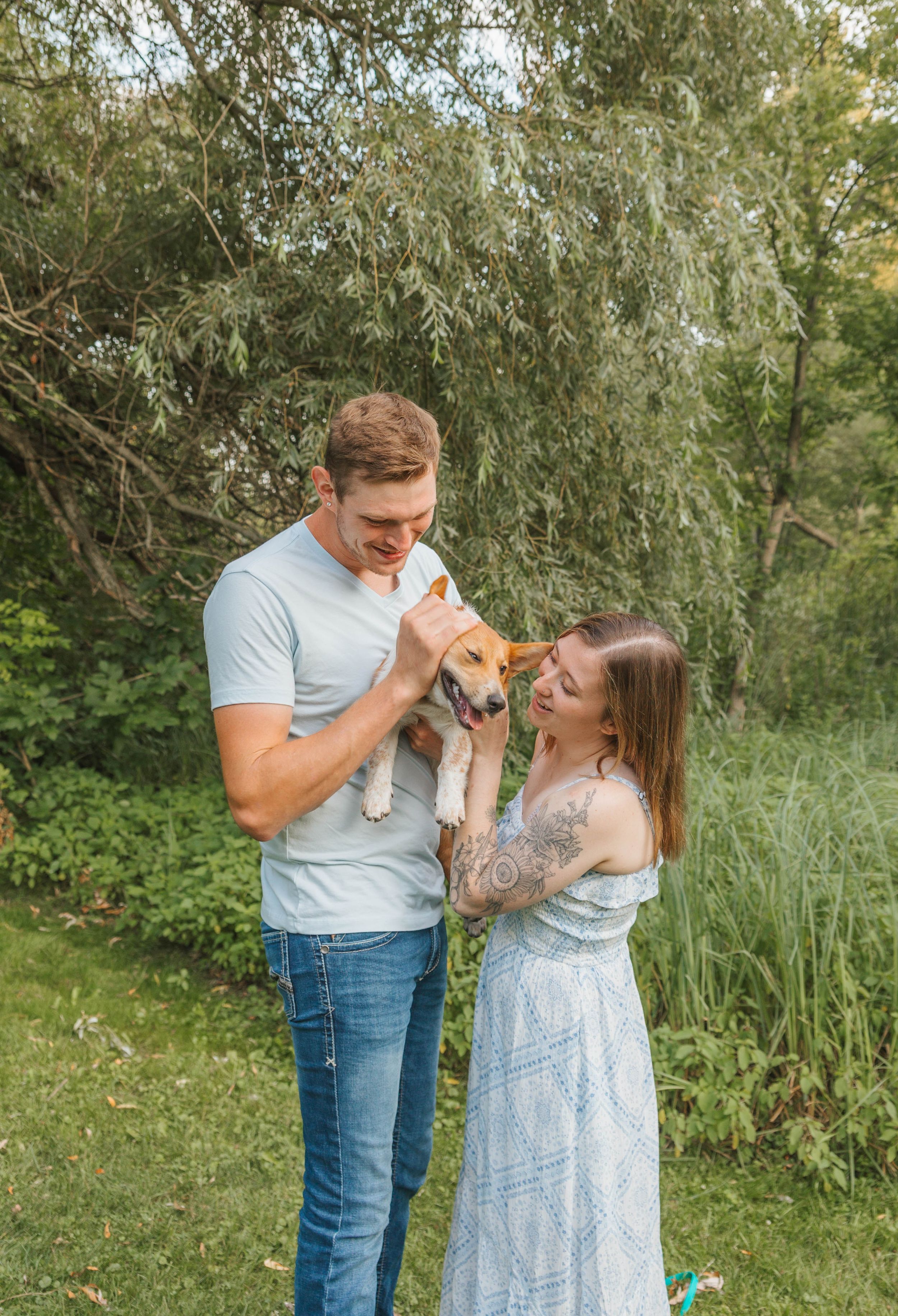 A smiling couple holds a small beagle puppy outdoors surrounded by lush green trees and tall grass.