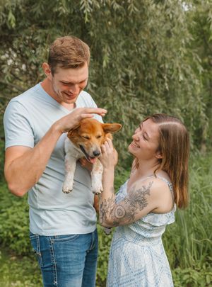 A smiling couple holds an adorable brown and white puppy outdoors surrounded by lush green foliage.