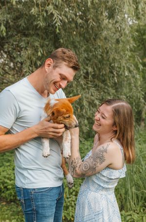 Happy couple smiling and holding an adorable brown puppy outdoors surrounded by lush green foliage.