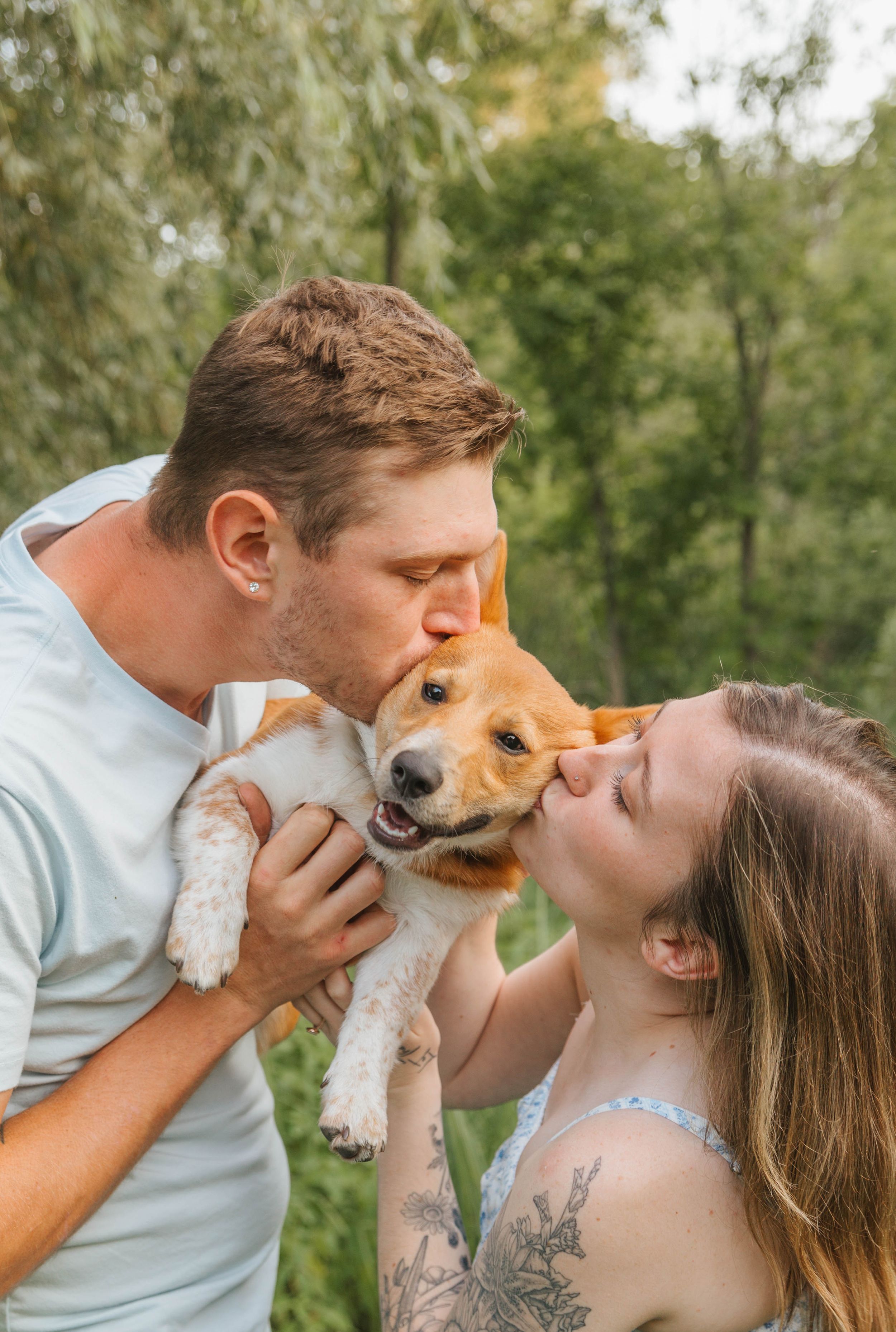 Couple kissing their happy Beagle puppy outdoors surrounded by lush green trees in a sweet pet photo.