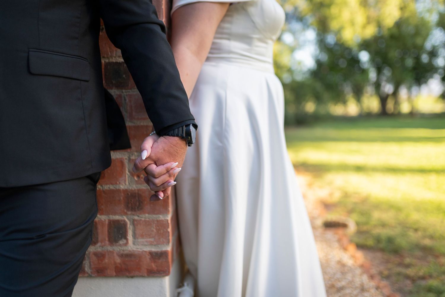 Bride and groom holding hands near brick wall outdoors on wedding day, white bridal gown and black suit visible.