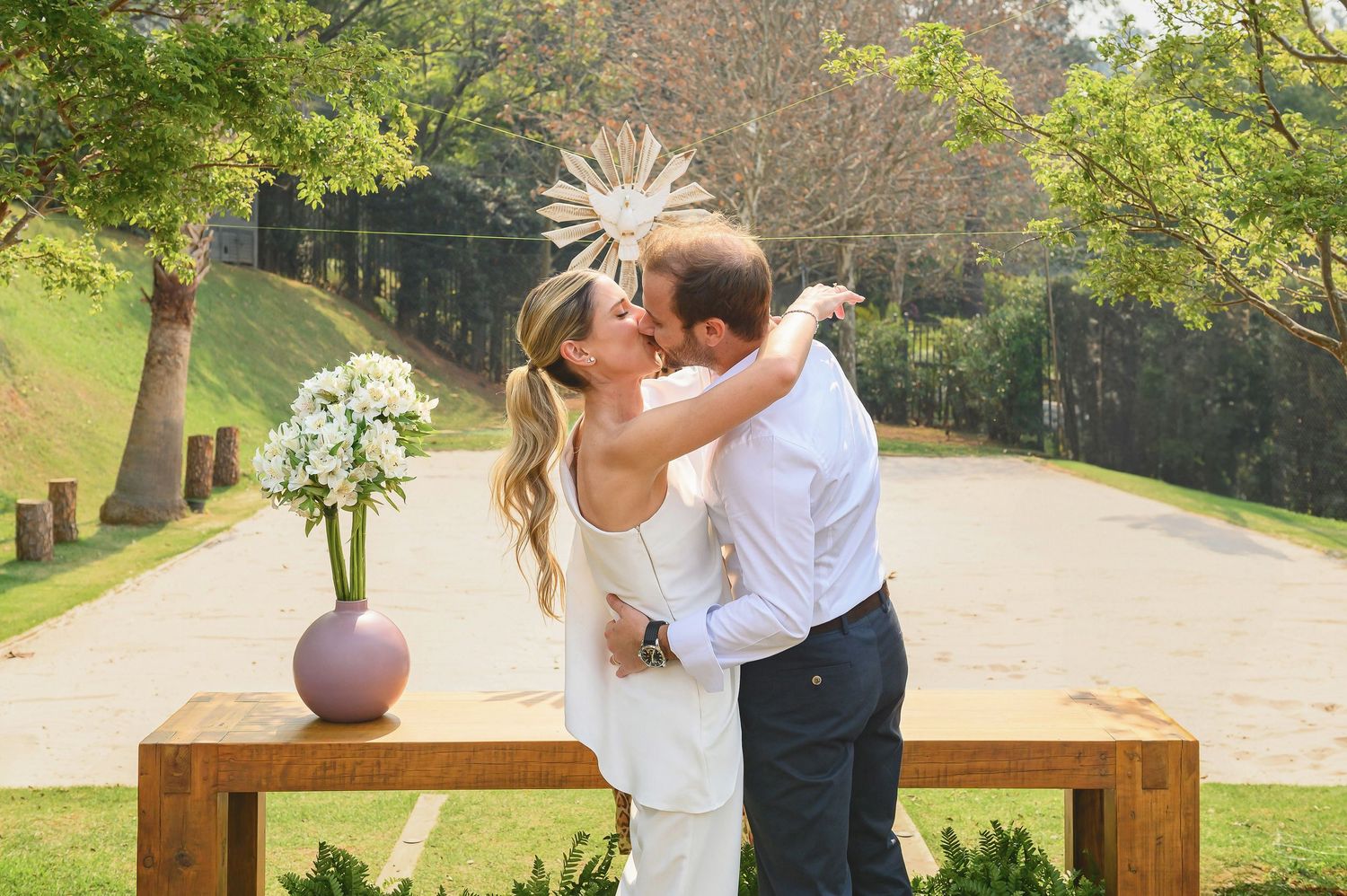 A bride and groom share a first kiss at an outdoor wedding ceremony with floral decor and lush greenery.