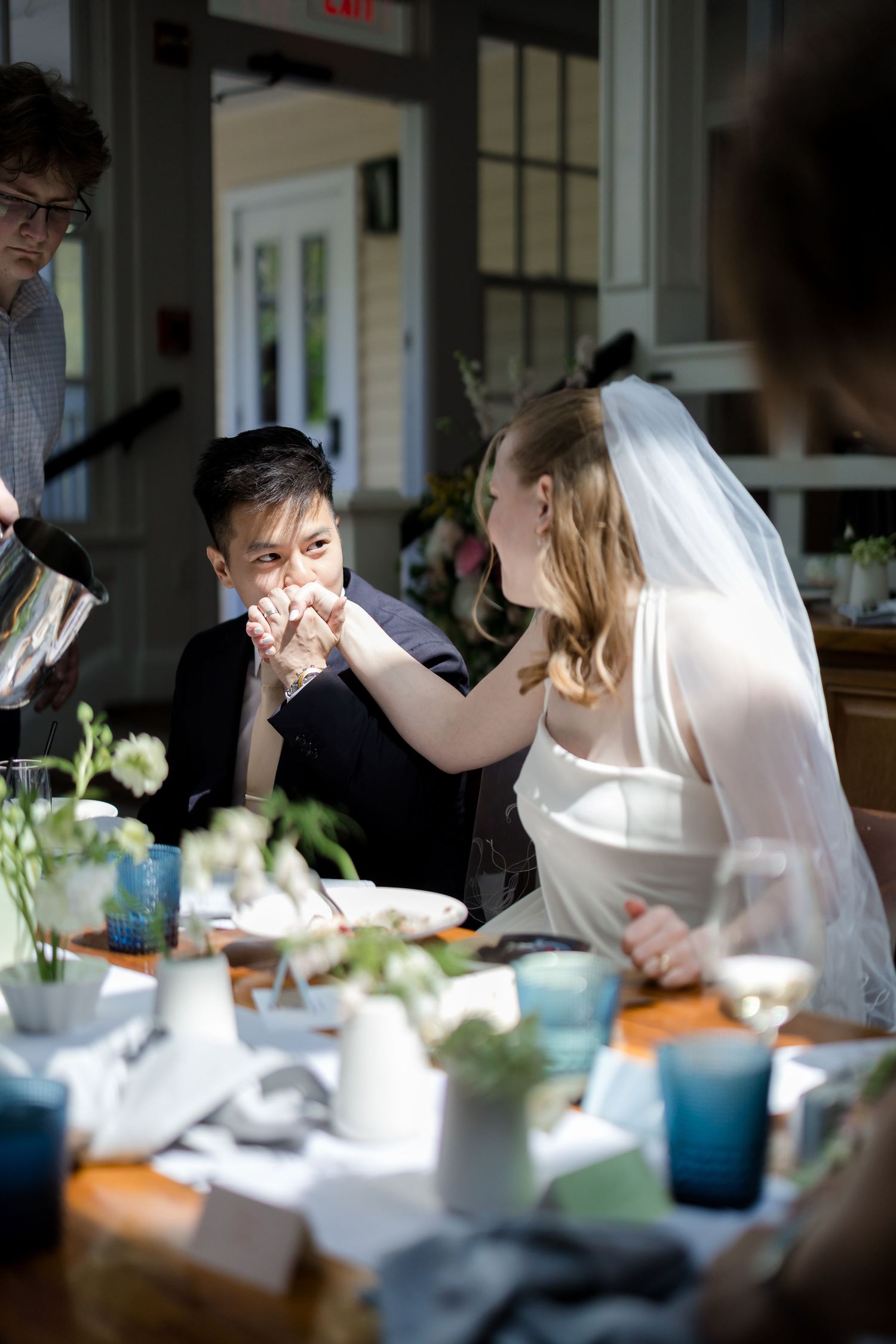 Bride in veil feeds groom cake at elegant wedding reception table with blue glassware and floral arrangements.