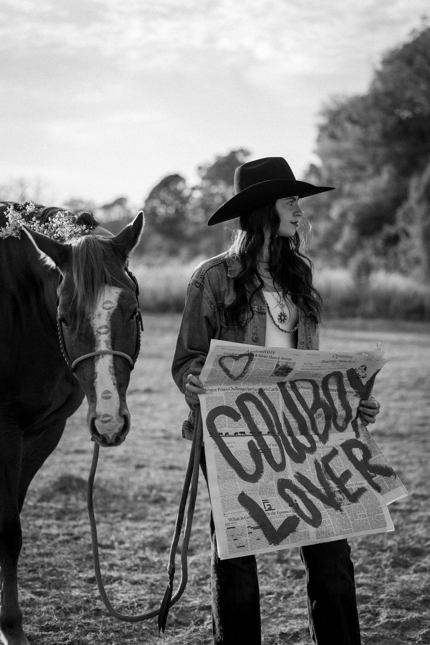 Woman in cowboy hat holding Cowboy Lover sign beside horse in field, black and white portrait.
