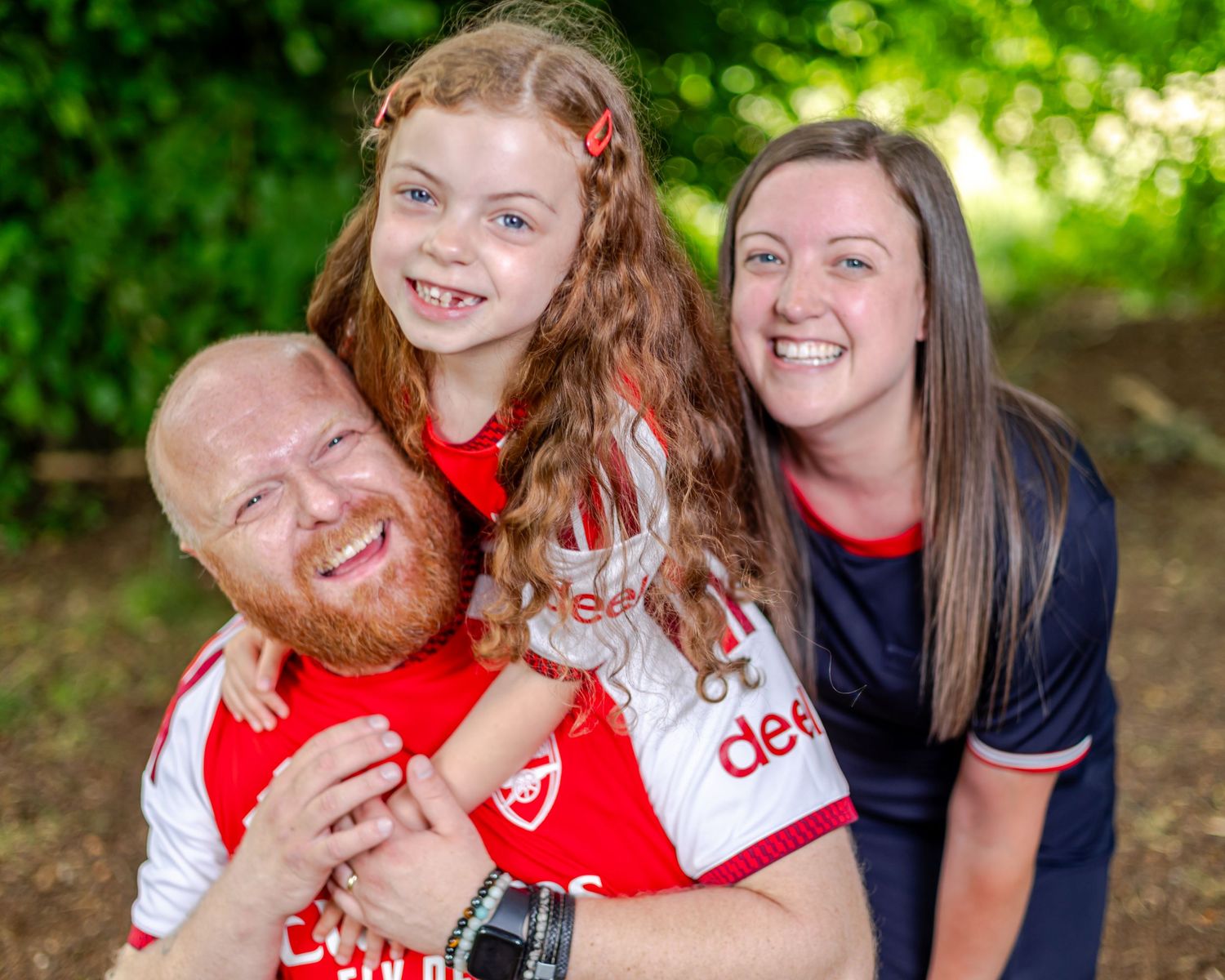 A smiling family of three in Arsenal jerseys pose outdoors, with a bearded dad holding his young red-haired daughter.