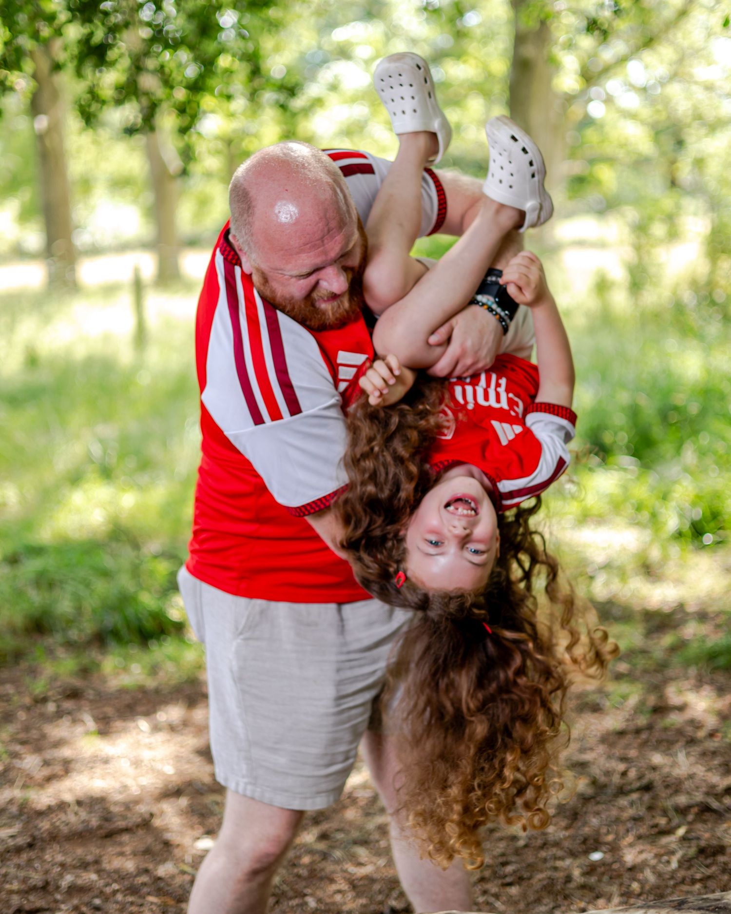 A bald bearded man in a red Adidas jersey playfully holds a laughing curly-haired child upside down outdoors.