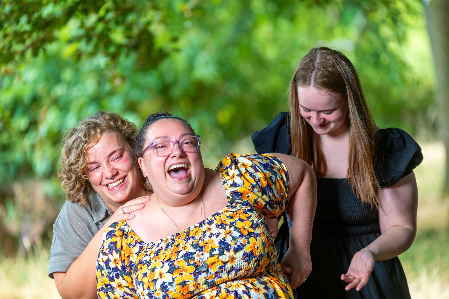 Three women laughing joyfully outdoors, with green trees in the background, sharing a warm and happy moment together.
