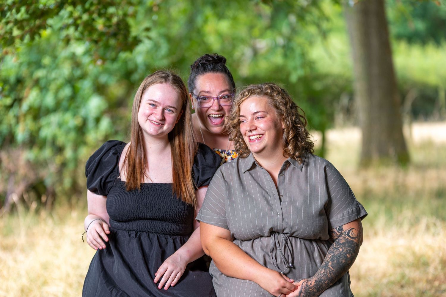 Three women smiling and posing together outdoors in a sunny park with lush green trees in the background.