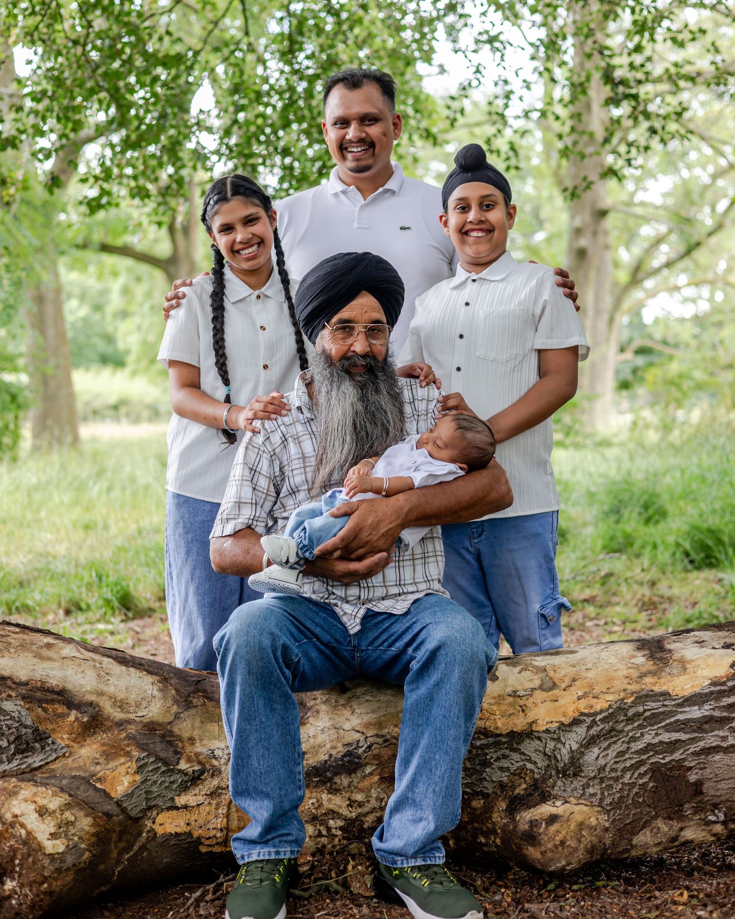 A multigenerational South Asian family poses outdoors on a log, with an elder holding a newborn baby surrounded by smiling relatives.