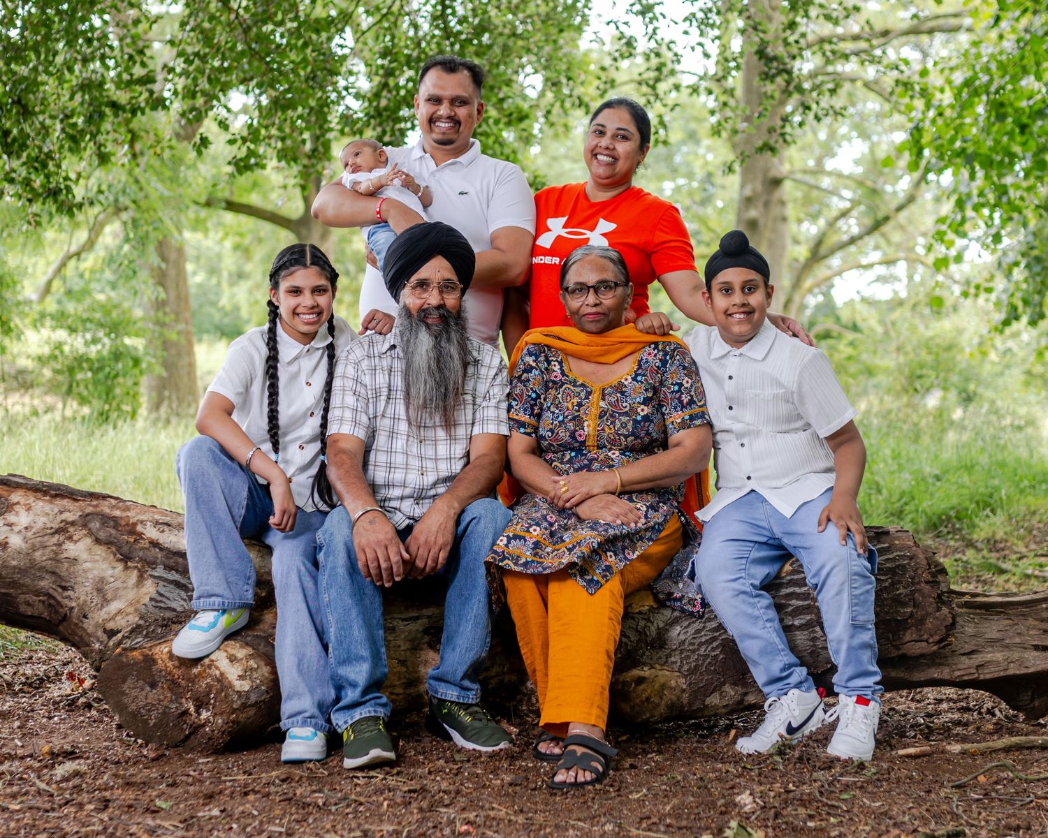 A Sikh family of seven poses together on a fallen log in a lush green park, smiling for a portrait.