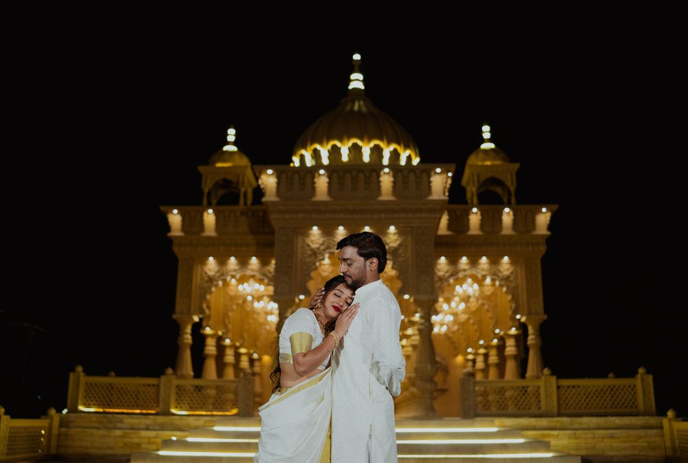 Couple in white attire embracing at night before an illuminated grand temple architecture.
