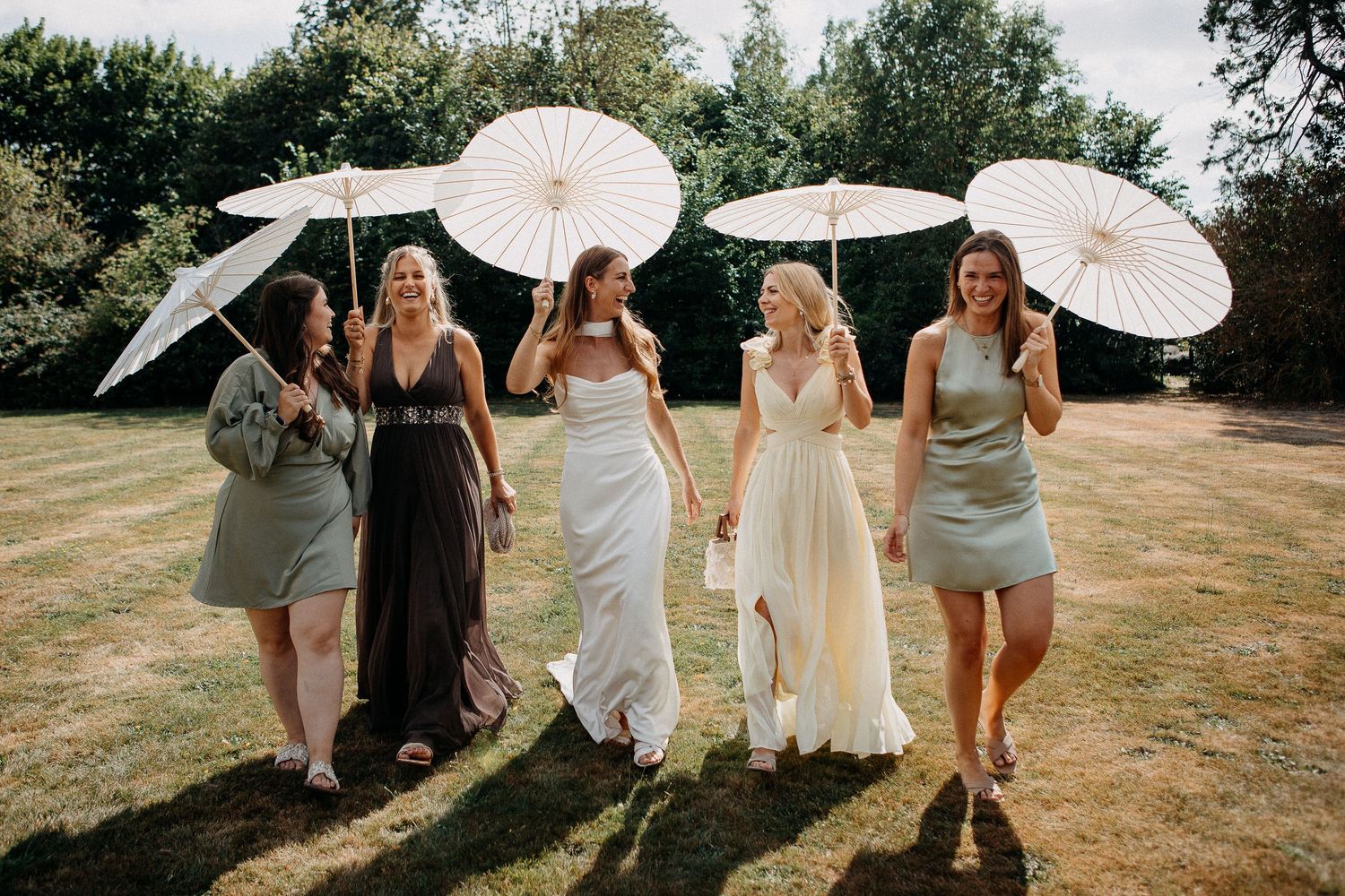 Bride and bridesmaids walking outdoors holding white parasols in summer garden wedding celebration.