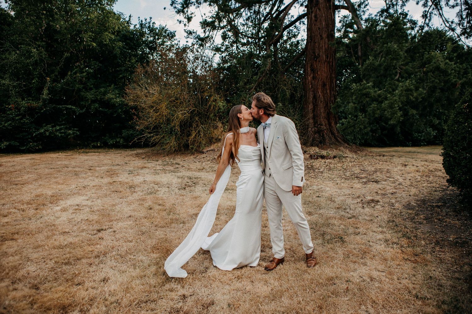Bride and groom sharing a kiss in a rustic outdoor setting with dry grass and tall trees.