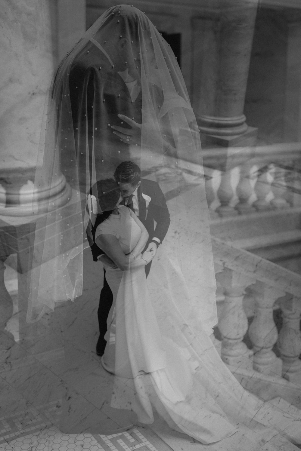 Black and white double exposure wedding photo of bride and groom kissing beneath flowing veil on elegant marble staircase.