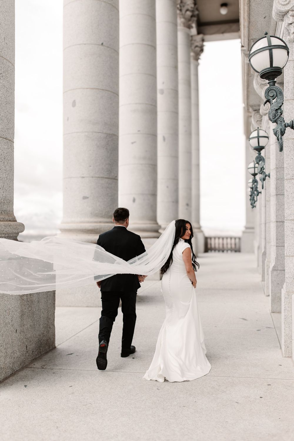 Bride and groom walking among grand white columns, her veil flowing in the wind, at an elegant wedding venue.