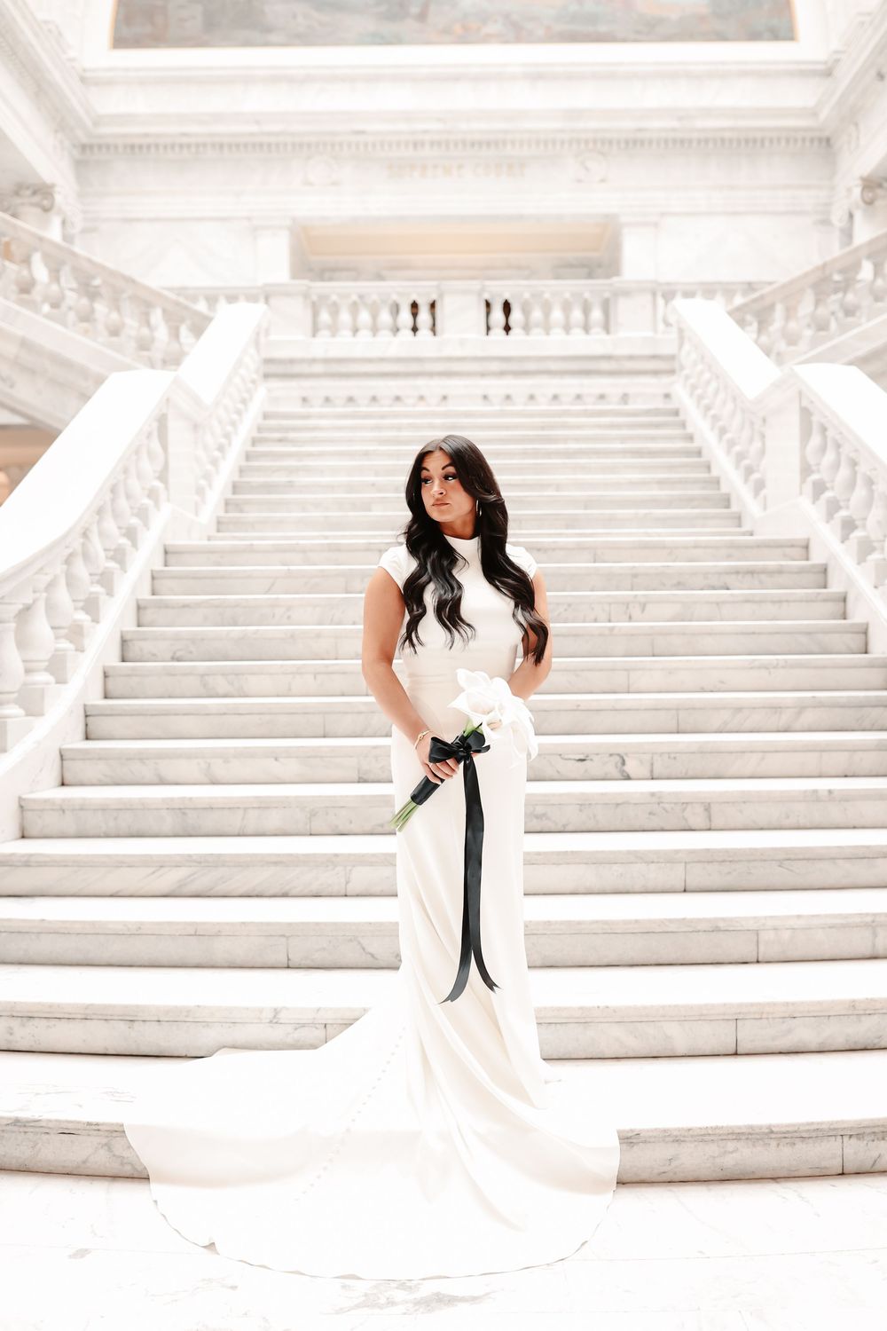 Bride in white mermaid gown with black sash holds bouquet on grand marble staircase.