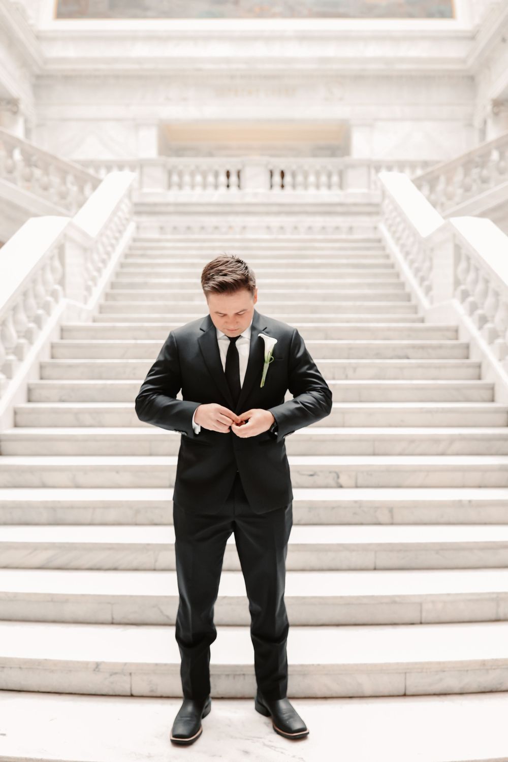 Groom in black suit with boutonniere buttoning jacket before grand marble staircase in elegant wedding venue.