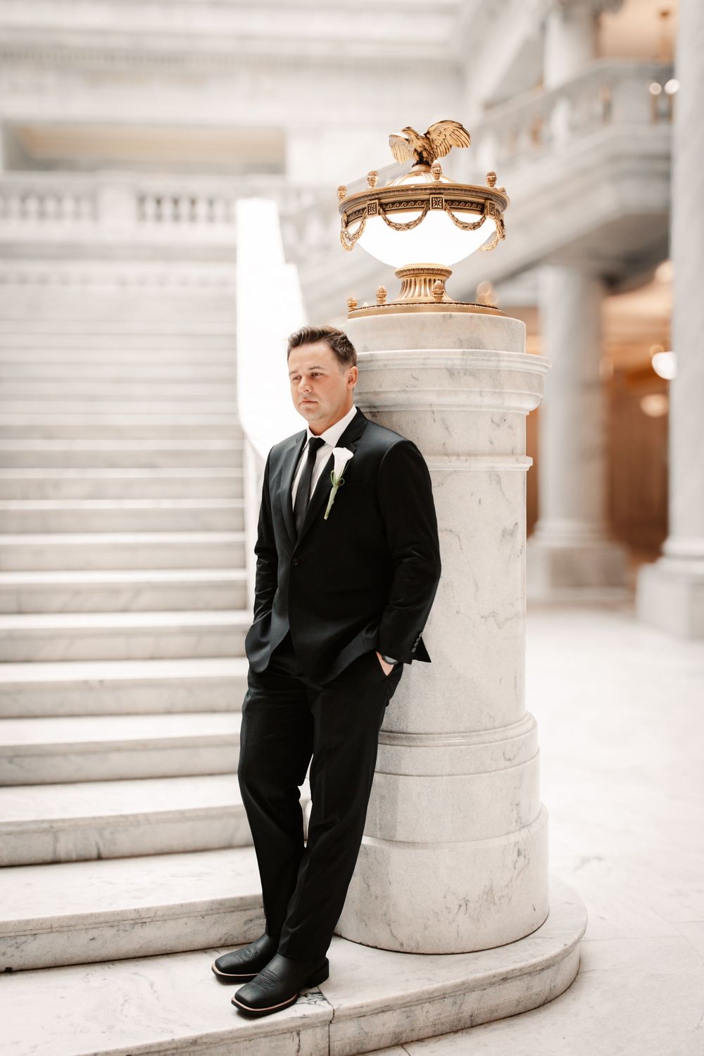 Groom in black suit leans against marble column in elegant neoclassical building with grand staircase.