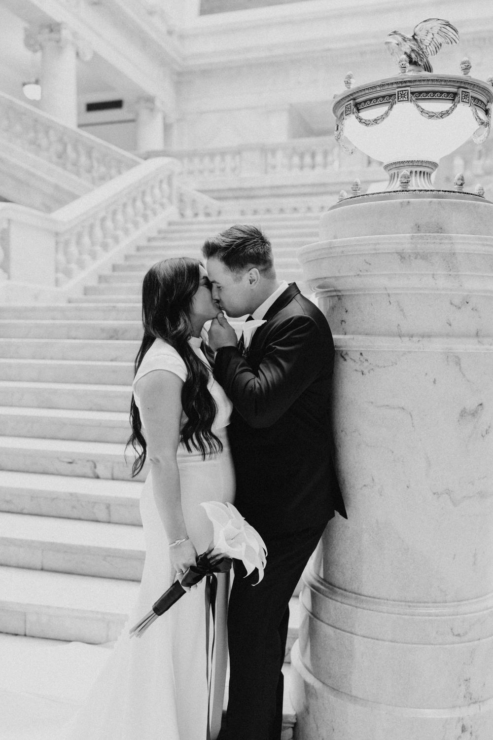 Bride and groom share a romantic kiss leaning against a marble column on an elegant staircase.