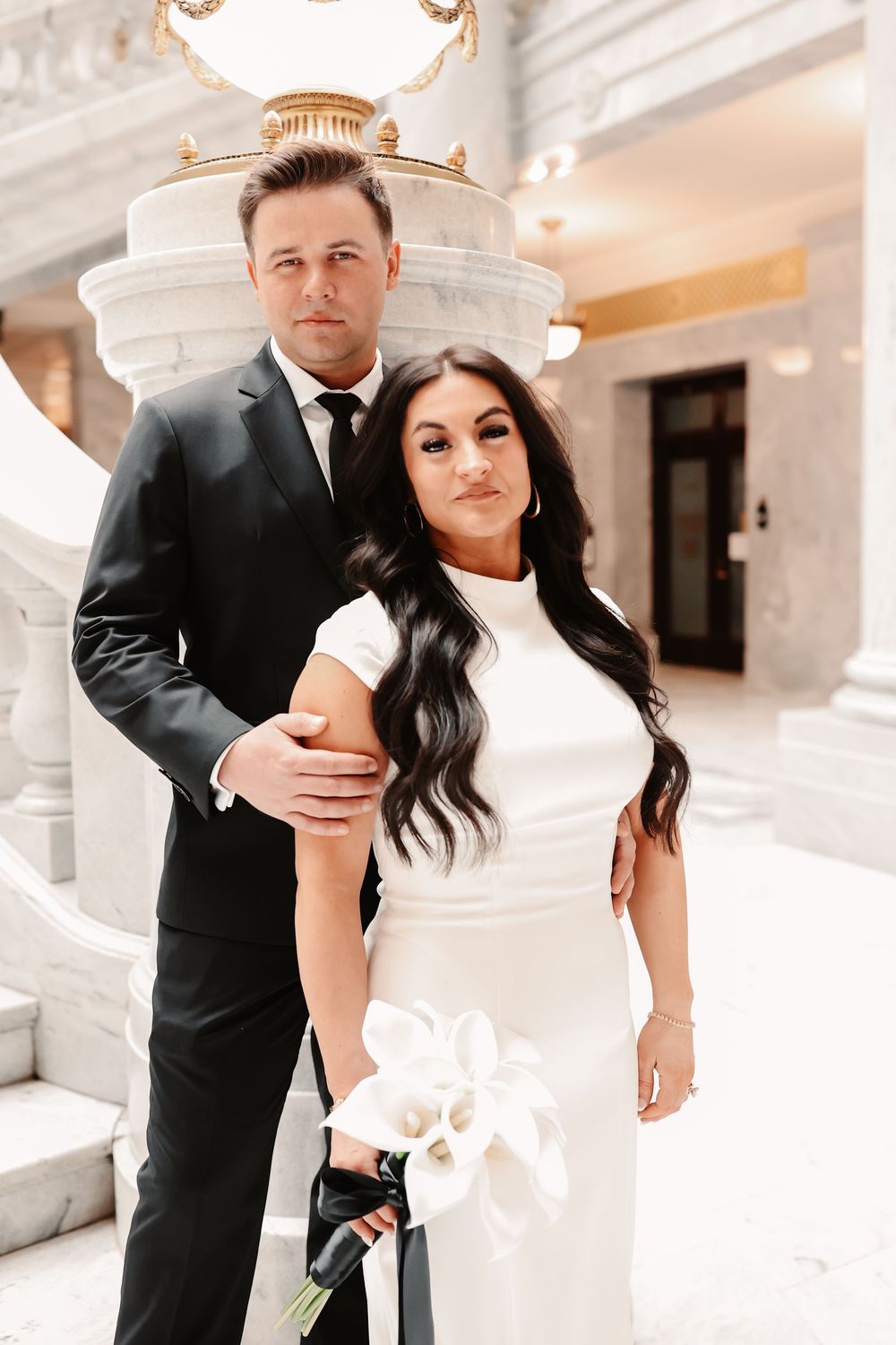 Elegant bride in white gown holding calla lilies stands with groom in black suit at a marble wedding venue.