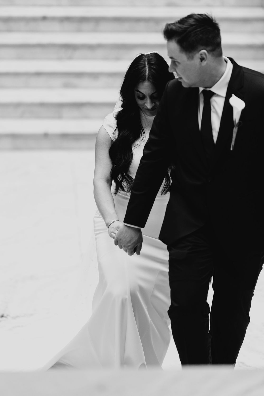 Black and white photo of bride and groom holding hands, walking together on steps in elegant wedding attire.