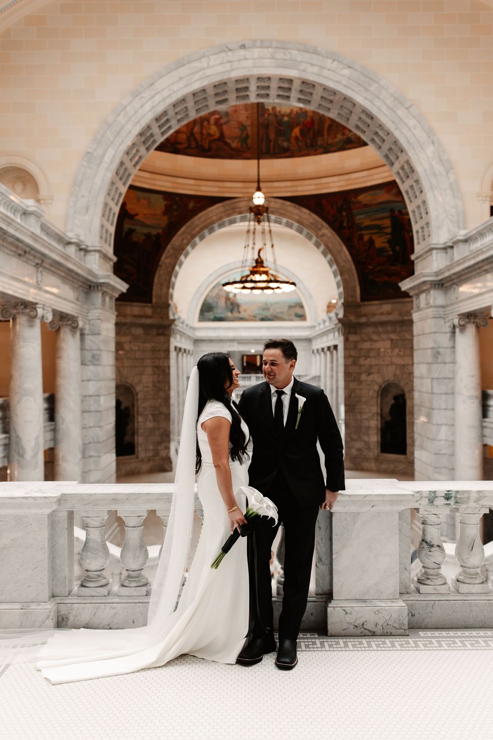 Bride and groom share a romantic moment inside an elegant marble building with grand arches and murals.