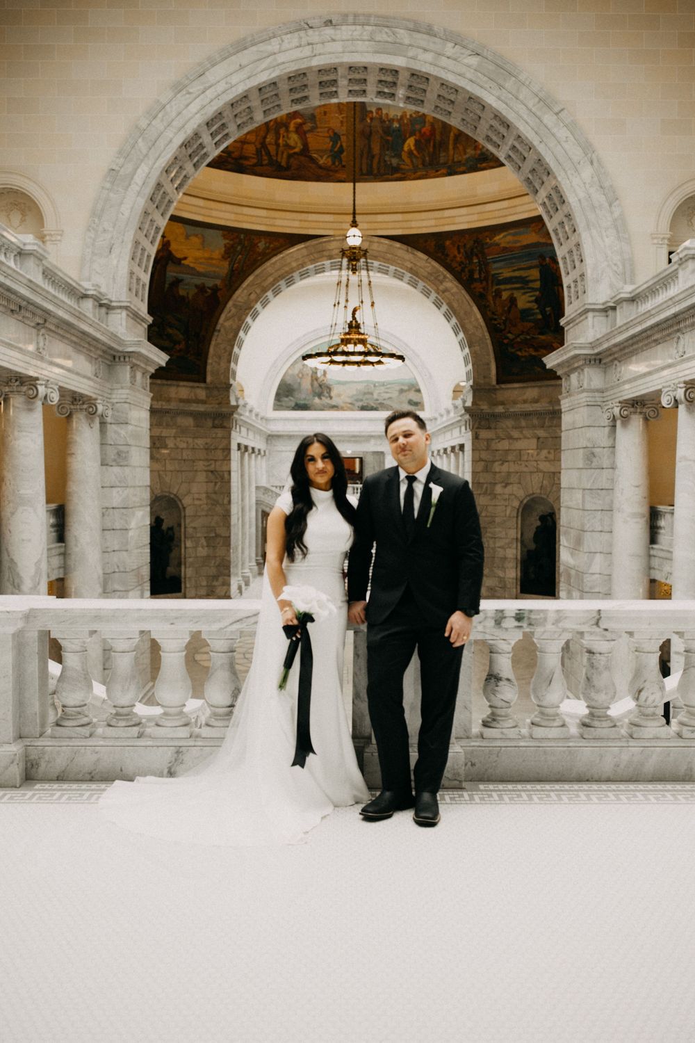 Bride in white gown and groom in black suit pose on marble balcony inside ornate capitol building.