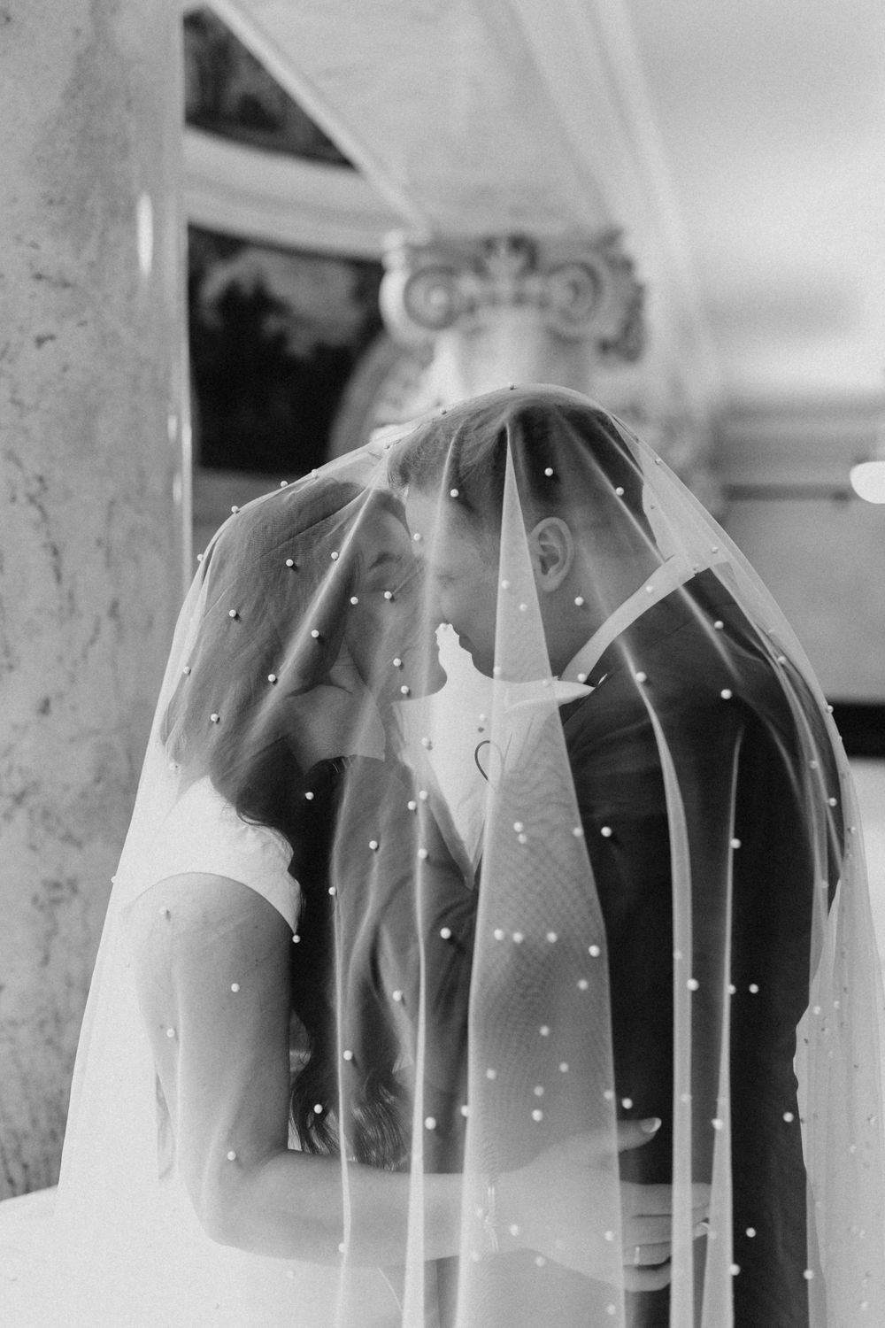 Bride and groom share intimate kiss under pearl-dotted wedding veil in black and white photo.