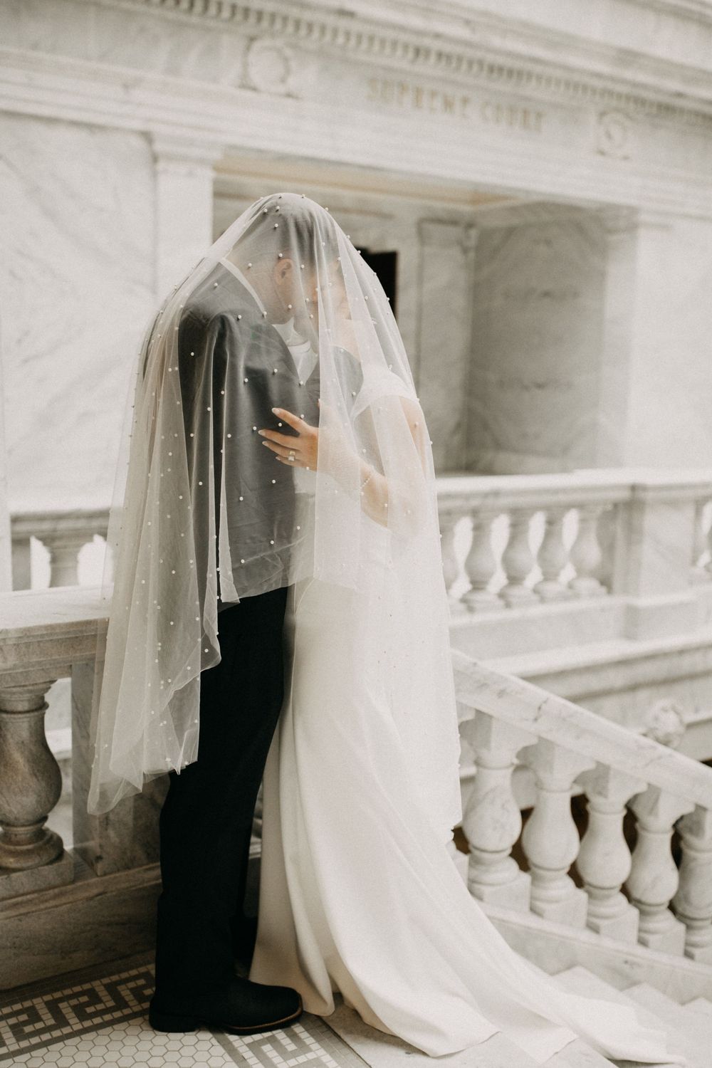Bride and groom kissing beneath flowing pearl-embellished veil on marble staircase at grand venue.