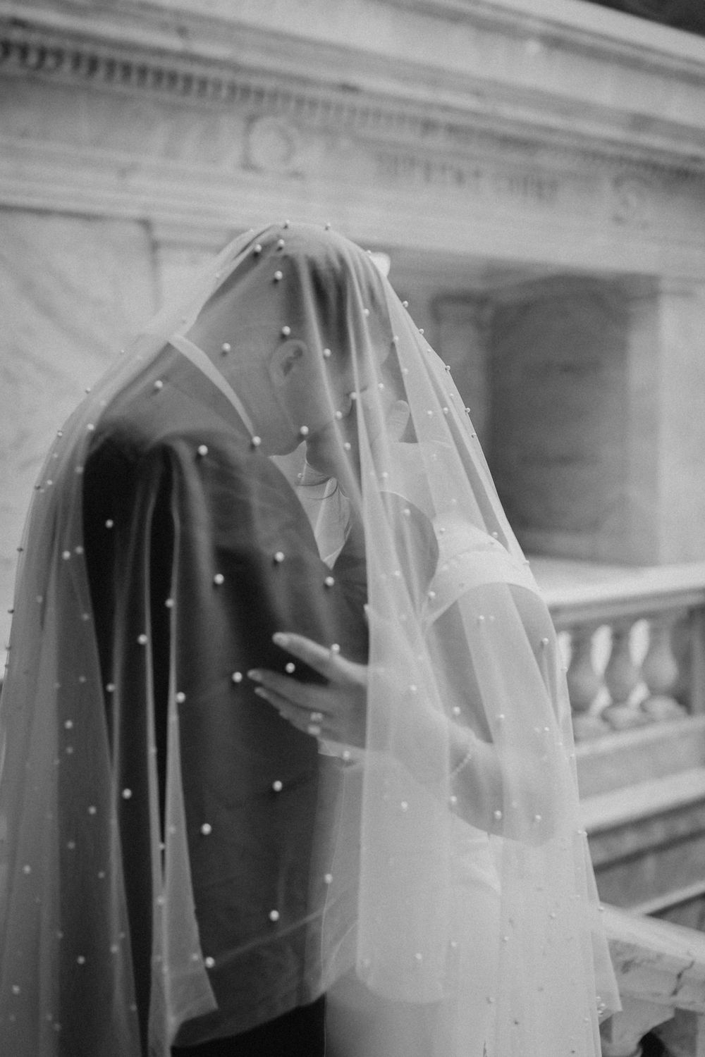 Black and white photo of couple embracing under sheer pearl-dotted bridal veil on marble balcony.