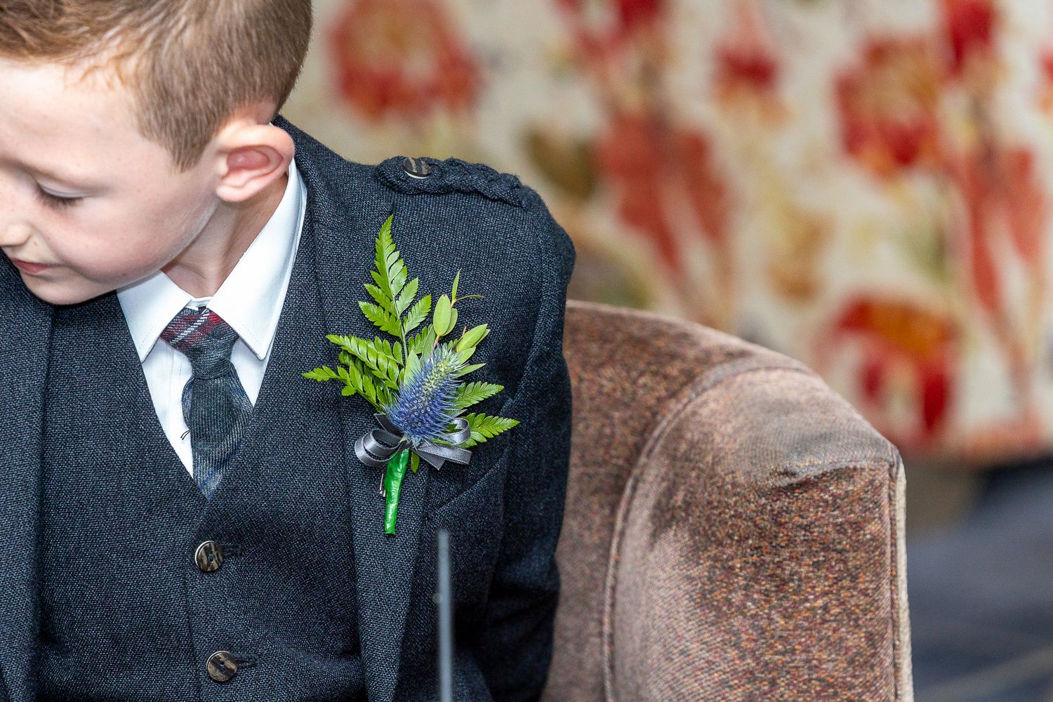 Young boy in dark suit with fern and thistle boutonniere at a formal event.