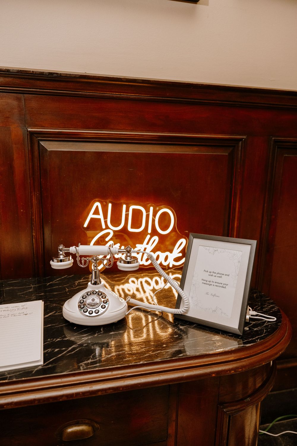 Vintage rotary phone audio guestbook station with glowing neon sign on dark wood console table at wedding reception.