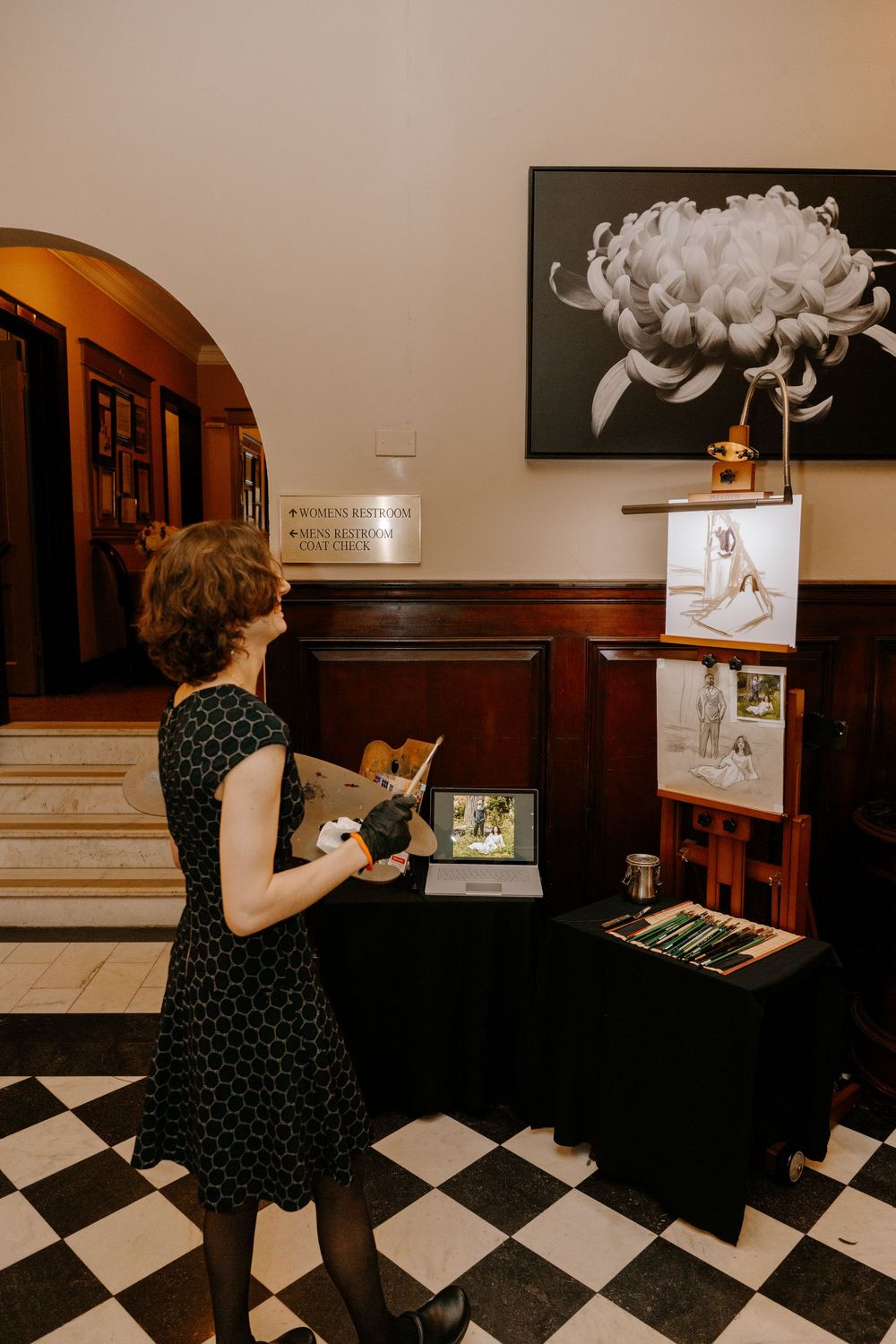 Woman in black dress viewing art display with tablet and drawings in elegant gallery with checkered floor.